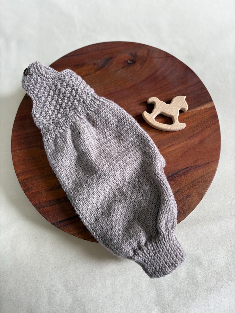 K&ouml;nnte beinhalten: Ein hellgrauer gestrickter Babysleeve mit strukturiertem Joch und Knopfverschluss. Ein kleines Holzschaukelpferd-Spielzeug befindet sich neben dem Sleeve. Der Sleeve liegt auf einem dunkelbraunen Holzbrett.