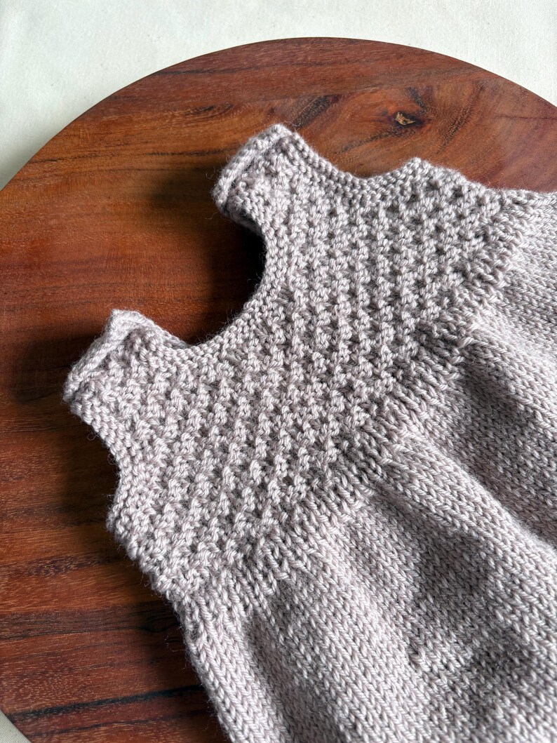 K&ouml;nnte beinhalten: Nahaufnahme eines hellgrauen Stricktops. Das Oberteil hat ein strukturiertes, gemustertes Joch und ein schlichtes, &auml;rmelloses Design. Das Kleidungsst&uuml;ck ist aus weichem Garn gefertigt und scheint handgefertigt zu sein.