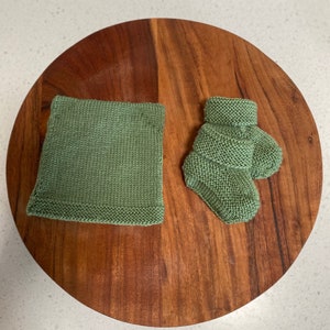 May include: A pair of green knitted baby booties and a green knitted square on a wooden surface.