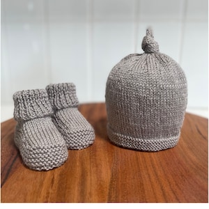 Puede incluir: Un conjunto de botines de bebé de punto gris y un gorro a juego con un nudo en la parte superior.
