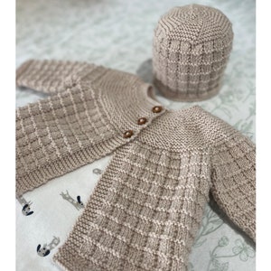 Könnte beinhalten: Ein beiger gestrickter Baby-Cardigan mit Knöpfen und einer passenden beigen Strickmütze.