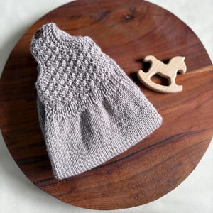 K&ouml;nnte beinhalten: Ein hellgraues gestricktes Babykleid mit strukturiertem Oberteil und Knopfverschluss. Ein kleines Holzpferd-Spielzeug liegt neben dem Kleid auf einer dunklen Holzoberfl&auml;che. Das Kleid ist wahrscheinlich handgefertigt.