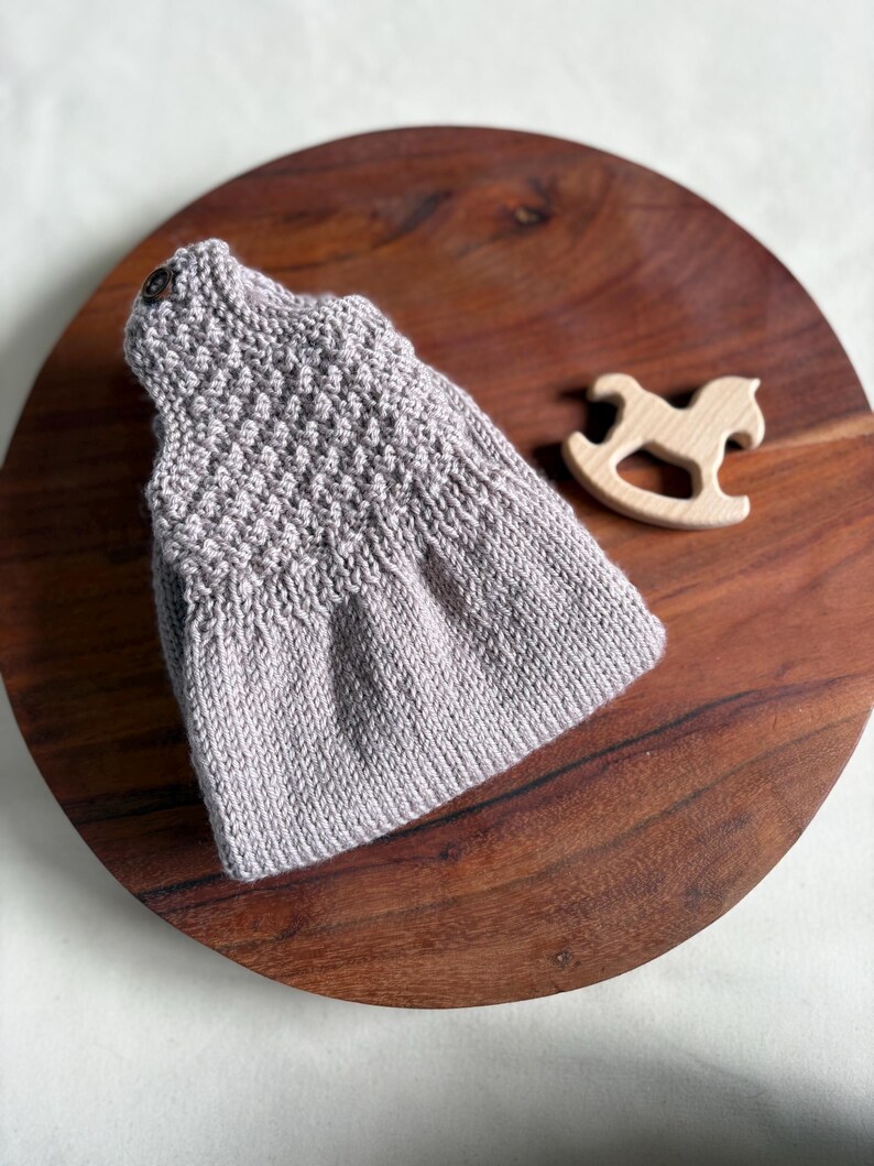 K&ouml;nnte beinhalten: Ein hellgraues gestricktes Babykleid mit strukturiertem Oberteil und Knopfverschluss. Ein kleines Holzpferd-Spielzeug liegt neben dem Kleid auf einer dunklen Holzoberfl&auml;che. Das Kleid ist wahrscheinlich handgefertigt.