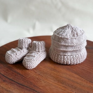 May include: A light gray knitted baby hat and matching booties set. The hat is a rounded shape with ribbed detailing. The booties have a ribbed cuff and textured pattern. The set is displayed on a wooden surface.