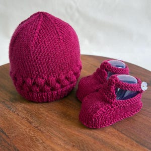 May include: A set of knitted baby booties and a matching hat in a deep burgundy color. The booties have a strap detail and a small white flower accent.