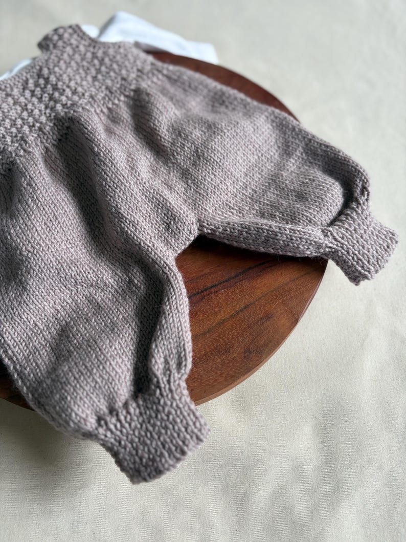 K&ouml;nnte beinhalten: Ein helllila Strickpullover mit strukturiertem Joch und gerippten B&uuml;ndchen. Der Pullover hat lange &Auml;rmel und wird auf einer Holzoberfl&auml;che pr&auml;sentiert. Das Kleidungsst&uuml;ck ist aus einem weichen, m&ouml;glicherweise wollenen Material gefertigt.