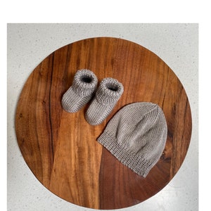 May include: A set of knitted baby booties and a matching beanie in a light brown color. The items are displayed on a wooden surface.