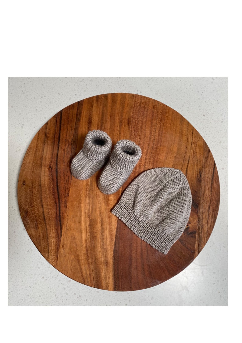 May include: A set of knitted baby booties and a matching beanie in a light brown color. The items are displayed on a wooden surface.