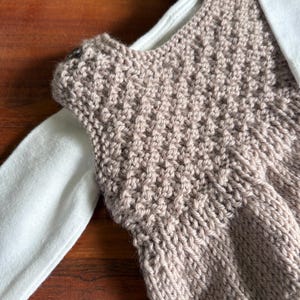 K&ouml;nnte beinhalten: Nahaufnahme eines gestrickten, &auml;rmellosen Oberteils in Hellbraun. Das Oberteil ist &uuml;ber einem wei&szlig;en Langarmshirt geschichtet. Das Strickwerk hat ein strukturiertes Muster, und die Gesamt&auml;sthetik ist handgemacht und gem&uuml;tlich.