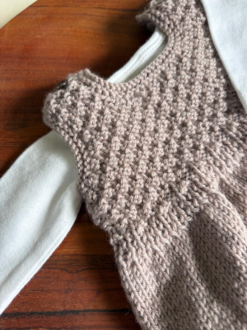 K&ouml;nnte beinhalten: Nahaufnahme eines gestrickten, &auml;rmellosen Oberteils in Hellbraun. Das Oberteil ist &uuml;ber einem wei&szlig;en Langarmshirt geschichtet. Das Strickwerk hat ein strukturiertes Muster, und die Gesamt&auml;sthetik ist handgemacht und gem&uuml;tlich.