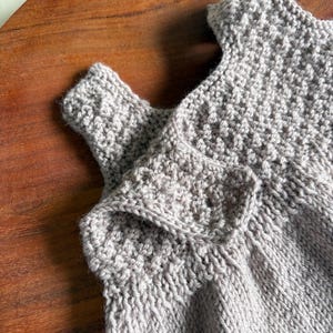 K&ouml;nnte beinhalten: Nahaufnahme eines hellgrauen gestrickten Babykleidungsst&uuml;cks. Das Kleidungsst&uuml;ck hat ein strukturiertes Strickmuster und einen Knopfverschluss. Der Artikel ist aus weichem Garn gefertigt und scheint handgefertigt zu sein. Der Hintergrund ist eine Holzoberfl&auml;che.