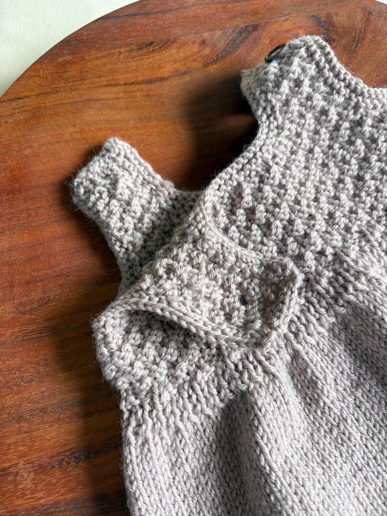 K&ouml;nnte beinhalten: Nahaufnahme eines hellgrauen gestrickten Babykleidungsst&uuml;cks. Das Kleidungsst&uuml;ck hat ein strukturiertes Strickmuster und einen Knopfverschluss. Der Artikel ist aus weichem Garn gefertigt und scheint handgefertigt zu sein. Der Hintergrund ist eine Holzoberfl&auml;che.
