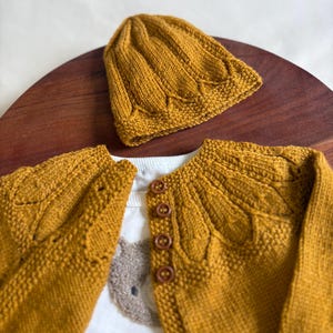 May include: Mustard yellow knitted baby cardigan and matching beanie hat. The cardigan has a button-up front and decorative leaf-like patterns. The hat has a ribbed design. A white shirt with a koala bear graphic is visible underneath.