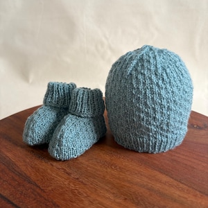 Pode incluir: Um conjunto de sapatinhos e chapéu para bebé na cor azul claro. Os sapatinhos e o chapéu são tricotados com um padrão texturizado.