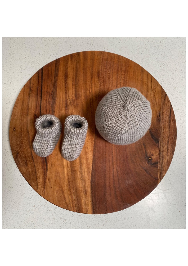 May include: A set of knitted baby booties and a matching hat in a light gray color. The items are displayed on a wooden surface.