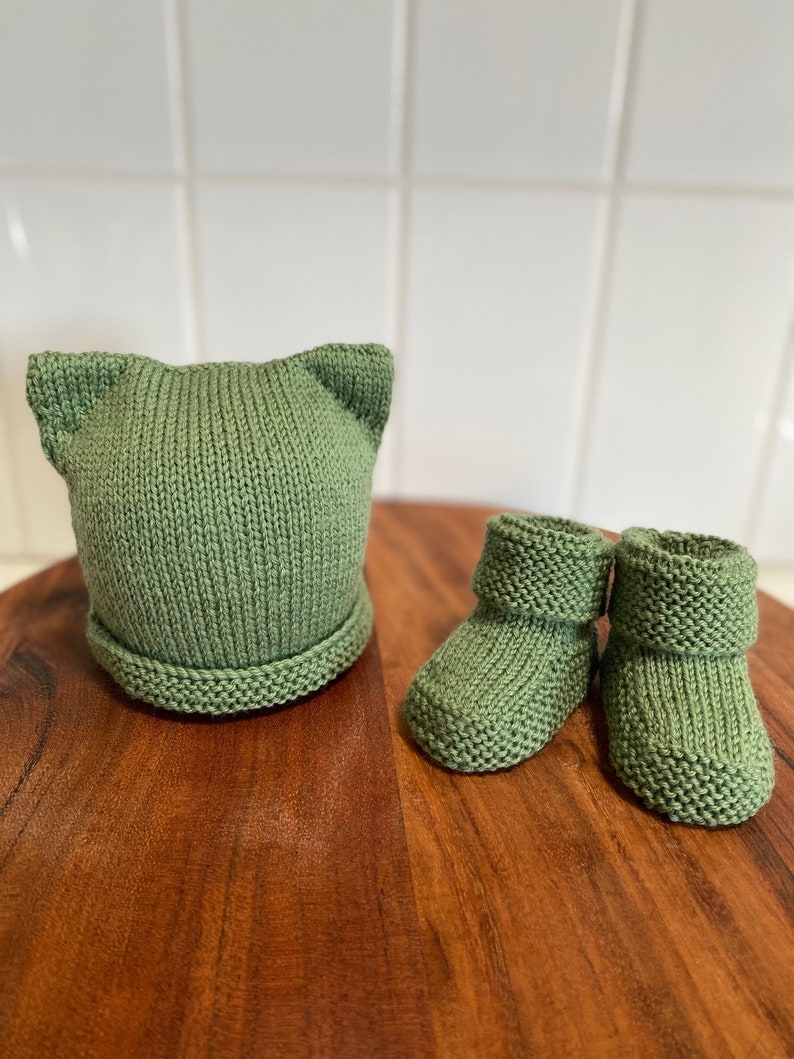 May include: A set of knitted baby booties and a hat in a sage green color. The hat has cat ears.