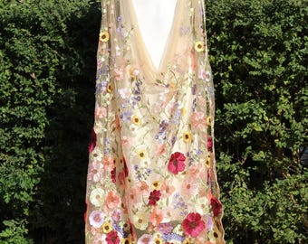 Pode incluir: Um xale transparente bordado com flores bege com um vestido branco por baixo. O xale é adornado com flores coloridas em tons de vermelho, rosa, amarelo e roxo. As flores estão agrupadas na parte inferior e espalhadas pelo tecido transparente.