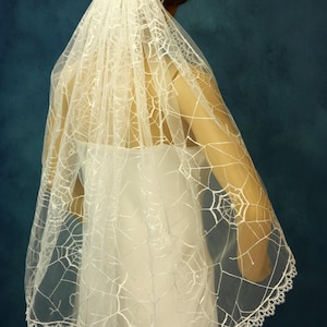 May include: A white sheer wedding veil featuring an embroidered spider web pattern across the fabric. The veil has a scalloped edge with a delicate lace trim and is draped over the shoulders, extending down the back in a layered design.