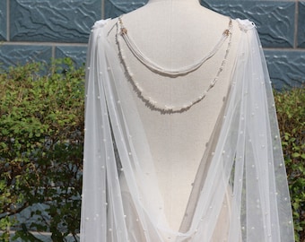 May include: A white tulle bridal cape with a beaded trim and a delicate pearl design. The cape is draped over a dress form and has a flowing, elegant look.
