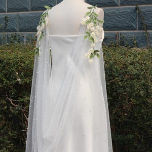 Könnte beinhalten: Eine weiße Tüll-Brautjacke mit Perlenbesatz und Blumenstickerei. Die Jacke hat ein fließendes Design und ist perfekt, um einem Brautkleid einen Hauch von Eleganz zu verleihen.