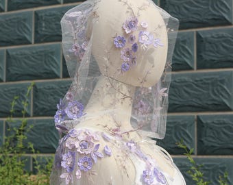 Pode incluir: Um véu transparente, cor marfim, adornado com delicadas flores de lavanda bordadas e vinhas. Os enfeites florais estão concentrados nas bordas do véu e descem em cascata pelo decote, criando um visual romântico e etéreo. O véu é exibido em um manequim.