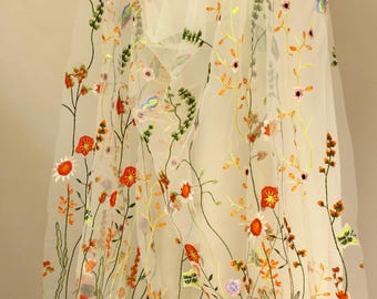 Pode incluir: Tecido branco transparente com bordados florais coloridos. O bordado apresenta flores laranja, vermelhas, amarelas e roxas com folhas e caules verdes. O tecido está drapeado, mostrando o design delicado.