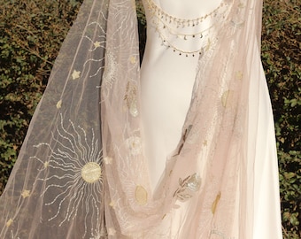 May include: A sheer, pale pink tulle veil featuring intricate gold and silver embroidery of suns, moons, and stars. The veil is draped over a plain white garment, which is adorned with a delicate gold chain necklace featuring small star and moon charms.