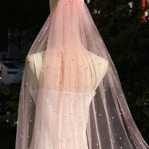 May include: A pale pink bridal veil adorned with small pearl beads. The veil is displayed on a mannequin, highlighting its fine, transparent material. The colour of the veil fades from a deeper pink at the top to a lighter hue at the bottom.