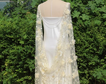 Pode incluir: Um vestido de noiva de cetim branco com uma sobreposição de renda transparente adornada com delicados detalhes florais. A sobreposição de renda tem um design fluido e drapeado e é acentuada por uma corrente prateada.