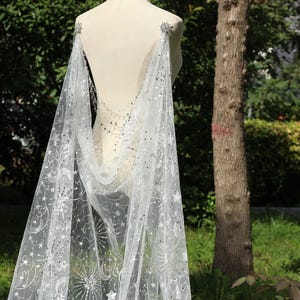 May include: A white, celestial-themed cape with star and moon patterns. The cape is draped over a mannequin, showcasing its flowing design. The fabric is adorned with silver embellishments, adding a touch of sparkle. Perfect for themed events.