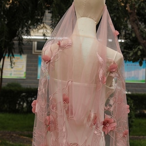 May include: A pale pink bridal veil with embroidered floral and leaf detailing. The veil is decorated with 3D fabric flowers and is draped over a mannequin, highlighting its design and texture. The background is blurred, with trees and a building visible.