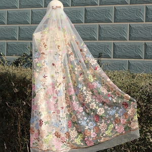 May include: An ivory-coloured veil with detailed floral embroidery in pink, blue, green, and brown. The veil is draped over a mannequin, highlighting the delicate floral design. The embroidery features a variety of flowers.