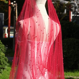 May include: A red tulle veil adorned with small, round beads. The veil is draped over a mannequin, highlighting its sheer, delicate fabric. The beads provide a subtle texture and visual appeal to the bridal accessory.