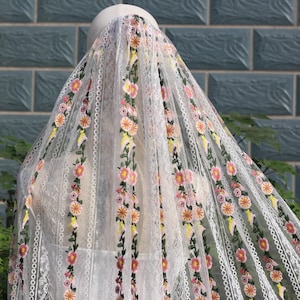 May include: A white bridal veil with floral embroidery in pink, yellow, and orange. The veil is draped over a mannequin, showcasing the delicate lace and intricate design. The background features a brick wall and greenery.