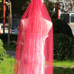 May include: A vibrant red, sheer fabric, adorned with small embellishments, is draped over a mannequin. The fabric is translucent, showing the form beneath. The background features greenery and a building.