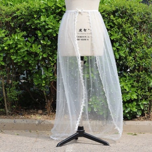 May include: A white tulle skirt with a pearl trim and a white satin ribbon tie. The skirt is on a white dress form.