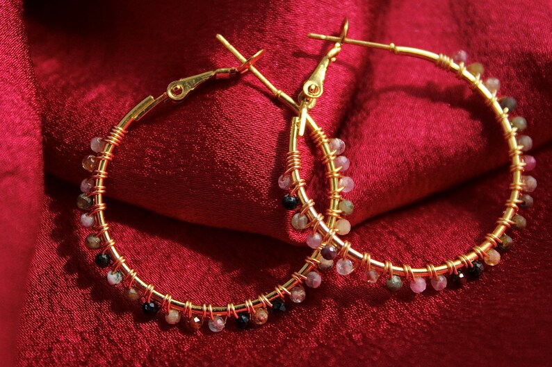 Peut inclure: Une paire de cr&eacute;oles dor&eacute;es avec un fil m&eacute;tallique enroul&eacute; le long des anneaux. De petites perles facett&eacute;es multicolores dans des tons de rose, noir et blanc translucide sont fix&eacute;es &agrave; intervalles r&eacute;guliers sur toute la circonf&eacute;rence.