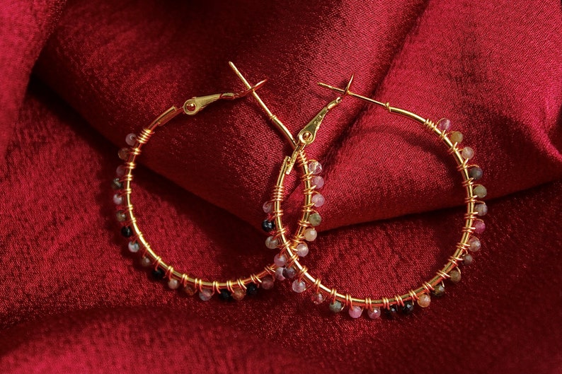 Peut inclure: Une paire de cr&eacute;oles dor&eacute;es avec un fil m&eacute;tallique fin enroul&eacute; autour des anneaux. De petites perles facett&eacute;es multicolores dans des tons de rose, vert et noir sont fix&eacute;es le long de la circonf&eacute;rence de chaque anneau avec du fil dor&eacute;.