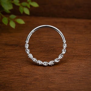 May include: A circular silver hoop ring featuring a twisted rope-like texture along the band. The jewelry piece has a smooth hinge mechanism at the top, allowing the hoop to open and close securely. The metal surface has a polished, reflective finish.