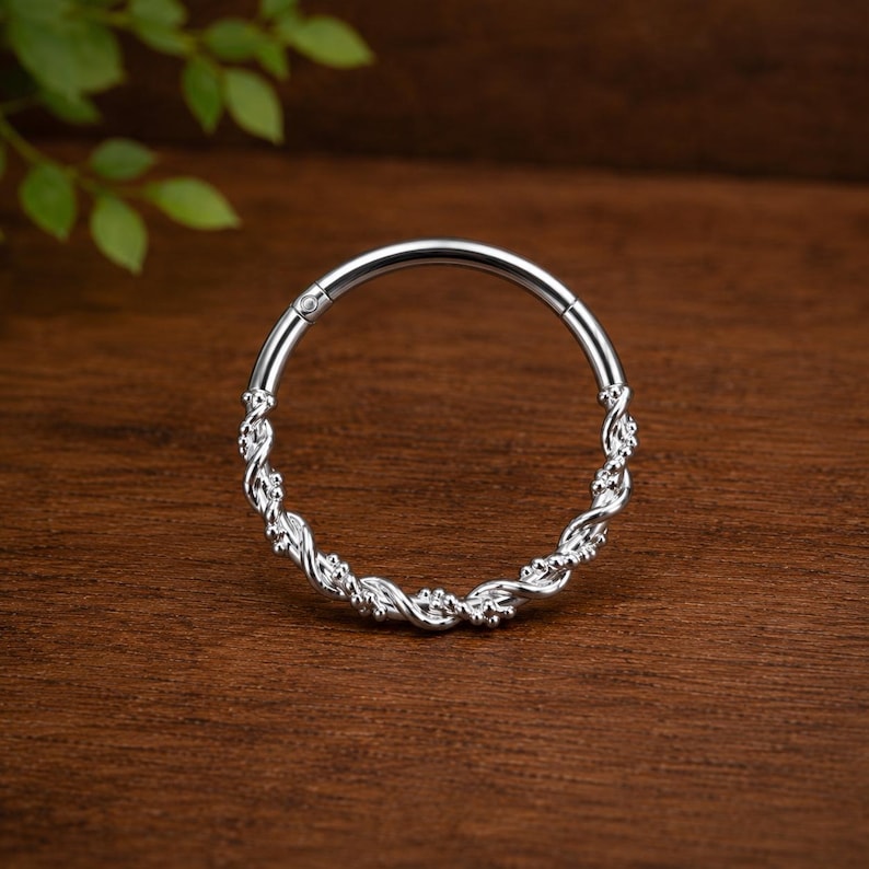 May include: A circular silver hoop ring featuring a twisted rope-like texture along the band. The jewelry piece has a smooth hinge mechanism at the top, allowing the hoop to open and close securely. The metal surface has a polished, reflective finish.