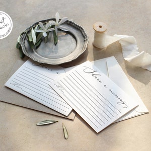 May include: Two white cards with black lines and the text "Share a memory" written in cursive. The cards are on a brown surface with a silver plate, white flowers, and a spool of white ribbon.