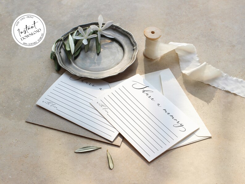 May include: Two white cards with black lines and the text "Share a memory" written in cursive. The cards are on a brown surface with a silver plate, white flowers, and a spool of white ribbon.