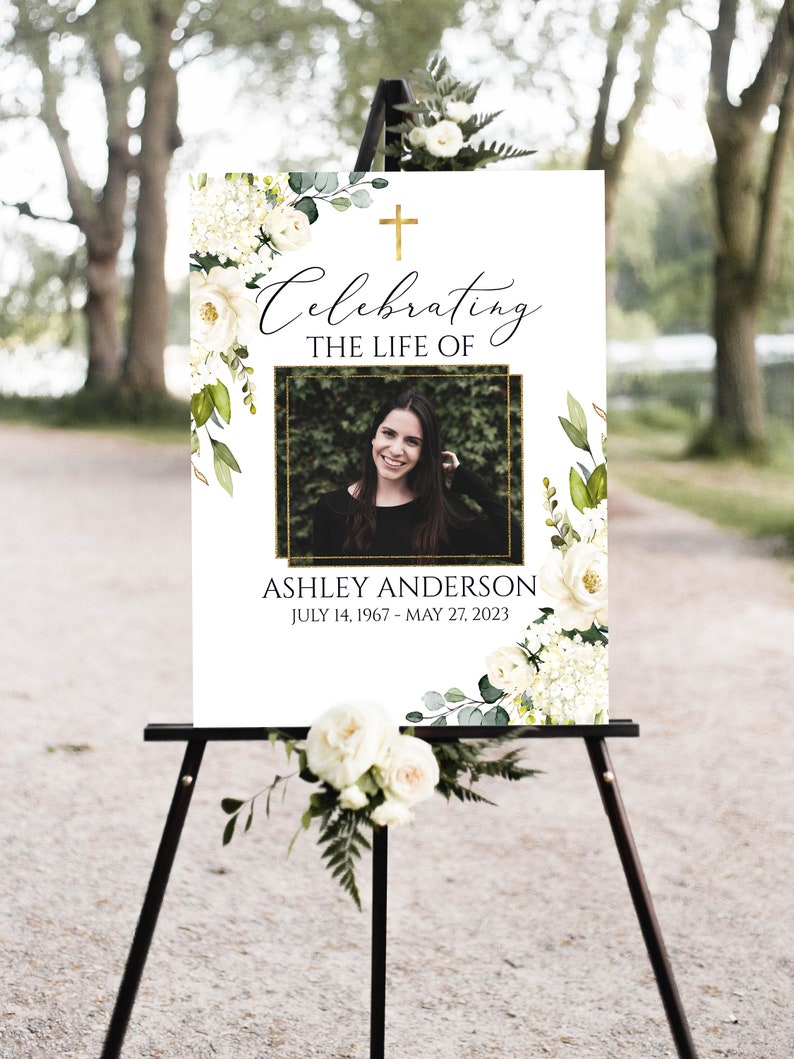 Pu&ograve; includere: Annuncio funebre floreale bianco con una croce dorata e una foto di una donna. Il testo recita "Celebrazione della vita di Ashley Anderson 14 luglio 1967 - 27 maggio 2023".