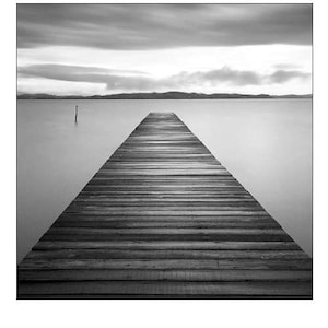 May include: A wooden pier extends out into a still, gray lake, with a distant shoreline and a cloudy sky.
