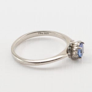 May include: A silver ring with a blue gemstone set in a halo of smaller stones. The ring is marked "D'Joy 925" on the inside band.
