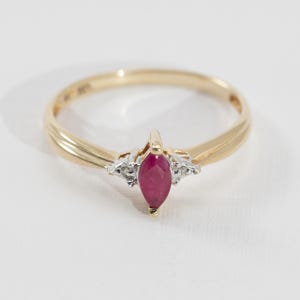 May include: A gold ring featuring a marquise-cut ruby gemstone as the centerpiece. The ruby is a deep red color, flanked by small, clear accent stones. The band is a polished gold with a simple, elegant design.