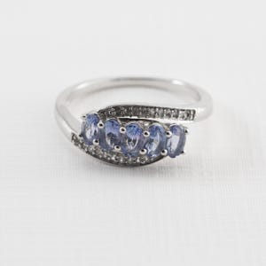 May include: A silver ring with a row of oval-shaped blue gemstones set in a curved band. The band is accented with smaller clear gemstones.