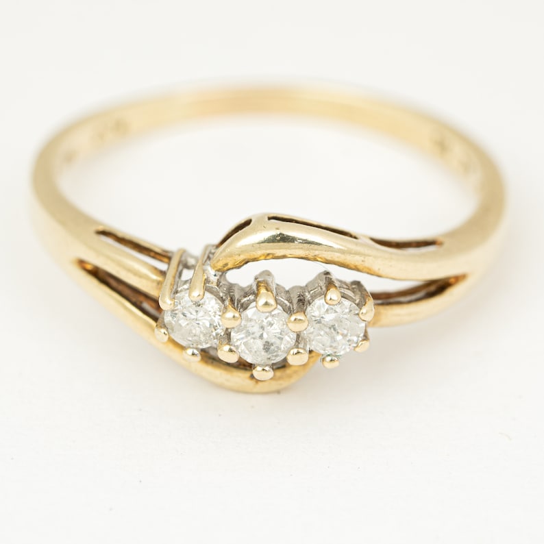 May include: A gold ring with three round, clear gemstones set in a row. The ring has a split shank design and a polished finish. The gemstones are set in a white metal, contrasting with the gold band.