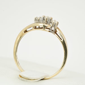 May include: A gold ring with a cluster of small diamonds. The ring has a curved design with a band that tapers towards the back. The diamonds are set in a cluster at the top of the ring. The ring is on a white background.