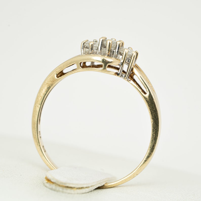 May include: A gold ring with a cluster of small diamonds. The ring has a curved design with a band that tapers towards the back. The diamonds are set in a cluster at the top of the ring. The ring is on a white background.